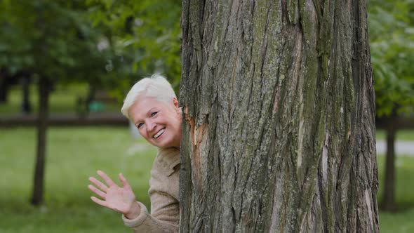 Cheerful Mature Middle Aged Woman Peeks Out From Behind Large Tree Plays Hide and Seek Smiling Older