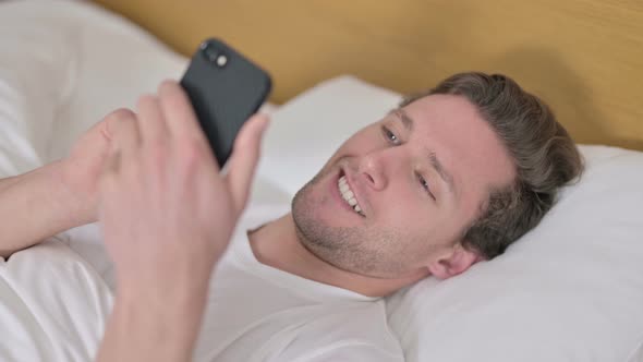 Portrait of Cheerful Young Man Using Smartphone in Bed