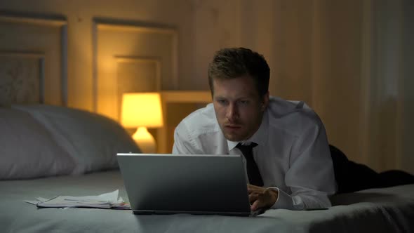 Thoughtful Male Lying on Bed and Reading Documents on Laptop, Business E-Mail