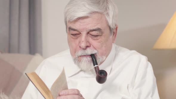 Close-up Face of Focused Old Caucasian Man with Smoking Pipe Reading Book. Confident Senior Man
