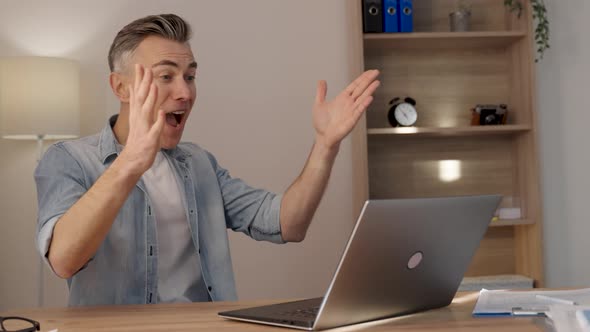 Caucasian man sitting at the table, looking at the laptop screen and rejoicing at good new