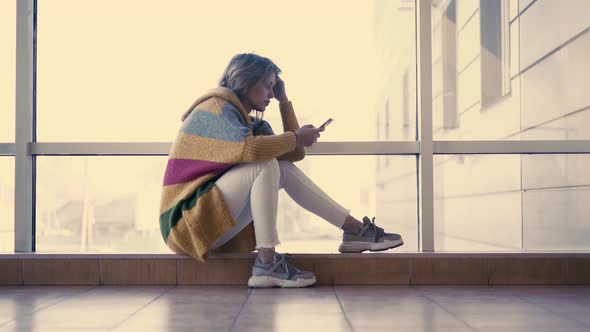 Young Cute Girl Sits on a Window with Smartphone in Her Hand Looking Thoughtfully at the Phone