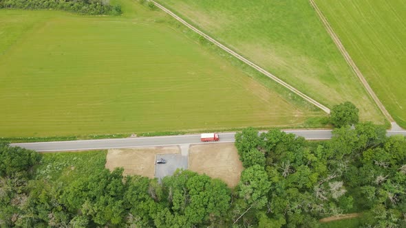 A red 5 ton truck driving along an empty curved road in between lush green meadows and thick leafy t