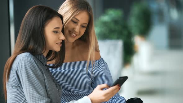 Two Attractive Young Women Holding Shopping Bags and Looking at Mobile Phone Together