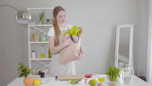 0Smiling Young Caucasian Woman Entering Kitchen with Bag of Products