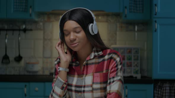 20s African American Woman Enjoys Listening the Music in Wireless Headphones Sitting at Kitchen at