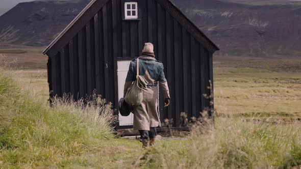 Stylish Man with a Sack Bag and a Banjo Case Walks Towards Little Black House