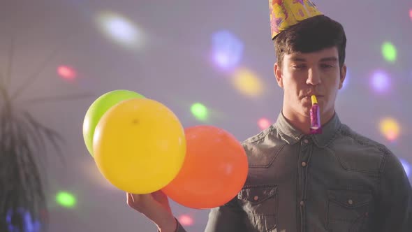 Portrait of Young Man in Birthday Hat with Noisemaker in Mouth Looking in the Camera Holding