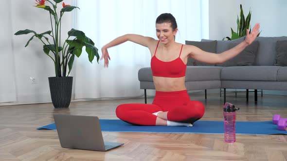 Smiling Woman in Sportswear Sitting on Yoga Mat Makes Wave with Hands at Home