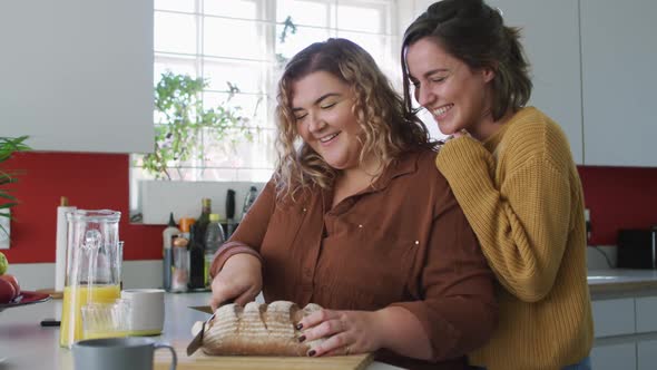 Happy caucasian lesbian couple slicing bread and laughing in kitchen