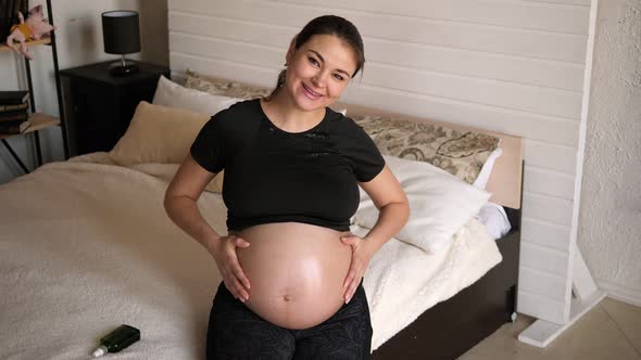 A Pregnant Woman Applies Moisturizing Oil To Her Stomach Sitting on the Bed