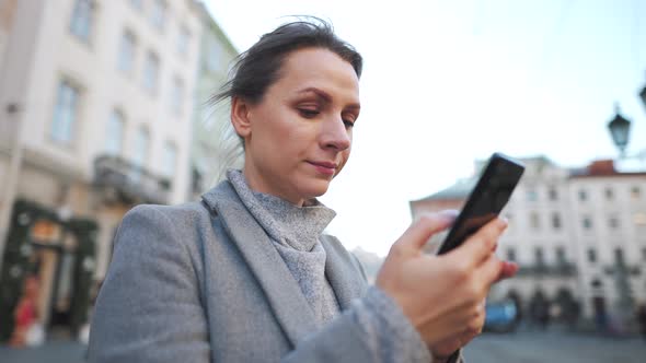 Woman in a Coat Standing in the Middle of an Old Square Using Smartphone and Taking a Photo