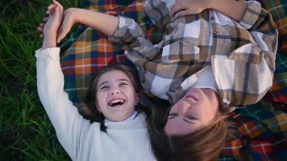 Young mum with a cute daughter lie on a blanket on a field at sunset