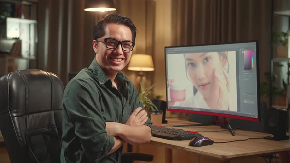 Asian Male Editor Works In Photo Editing Software. He Turns And Warmly Smiles Into The Camera