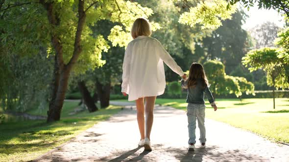 Back View Happy Young Mother and Little Daughter Holding Hands Walking in Sunrays in Slow Motion