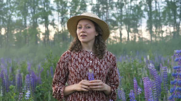 Romantic Feminine Woman in Hat and Dress Stands in a Flower Field in Fog and Holds Lupine in Hands