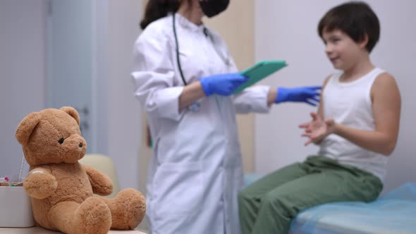 Closeup Brown Teddy Bear in Pediatric Clinic with Blurred Caucasian Boy Talking to Unrecognizable