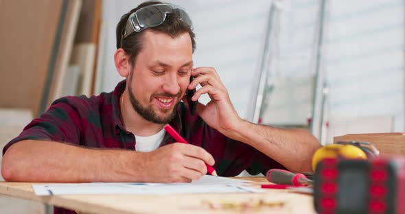 Portrait of Modern Young Bearded Man Speaking By Phone with Client Smiling and Making Notes in