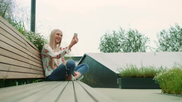 Lovely Young Female Using Smartphone To Make Video Call While Sitting on Bench on Living Roof of