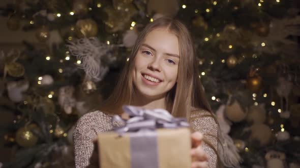 A Young Pretty Girl Smiles and Holds Out a Christmas Present To the Camera.