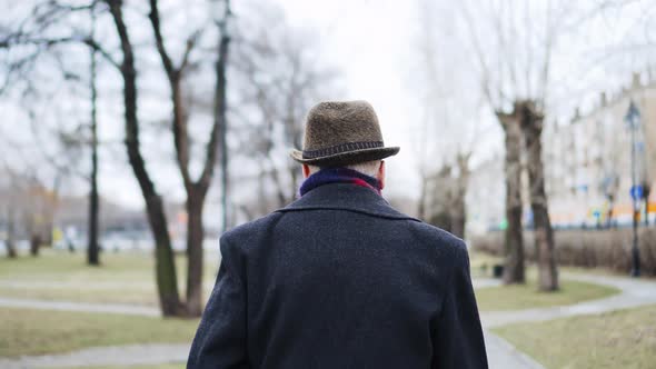 Old man in hat is walking in city park, back view. He limps on his leg, lame man.