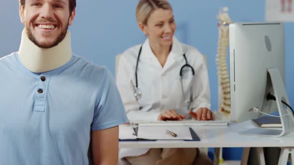 Patient smiling at camera while female physiotherapist working over computer in background