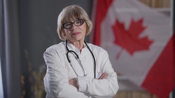 Confident Senior Woman in White Medical Gown Crossing Hands Looking at Camera with Canadian Flag at