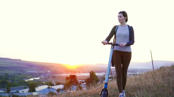 Young Girl Walks Down the Hill with an Electric Scooter and Watches the Sunset