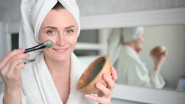 Beautiful Smiling Woman at Home in Bathrobe with a Towel Applying Face Clay Mask Against Acne To