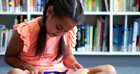 Schoolgirl sitting on floor and reading a book in library at school