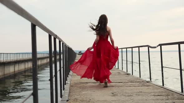 Young Lady with Curls in Scarlet Dress Runs Quickly Towards the End of the Pier