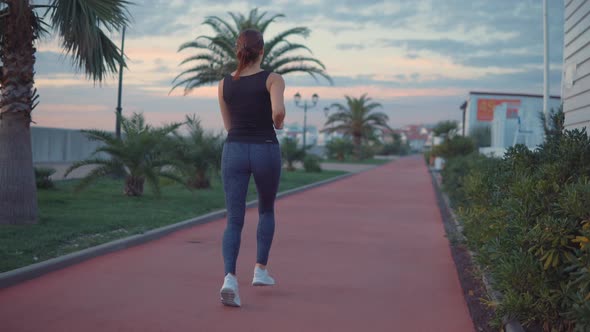 Slim Woman Is Jogging in City Embankment Along Small Houses, Keeping Fit