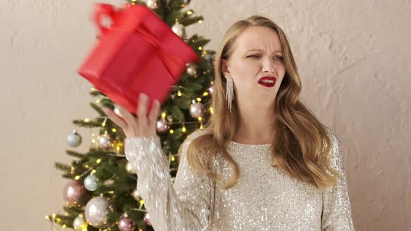 Festive woman in a shiny evening dress near Christmas tree gets a bad gift and throws it away