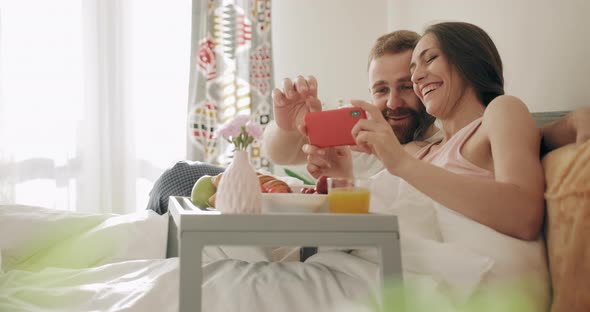 Cheerful Family Watching Photos While Sitting on Bed with Tray Full of Food. Young Woman and Man