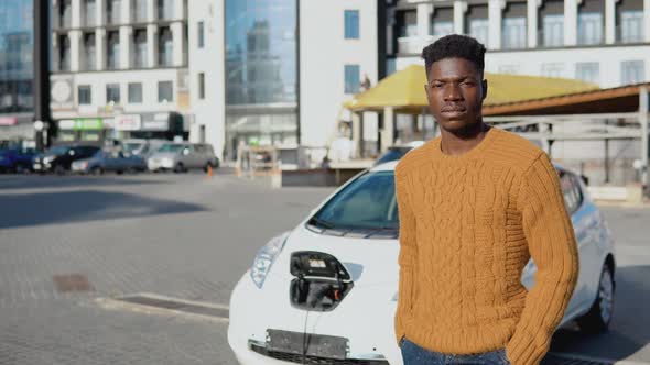 African American Driver is Standing Near a White Electric Car While Charging the Battery