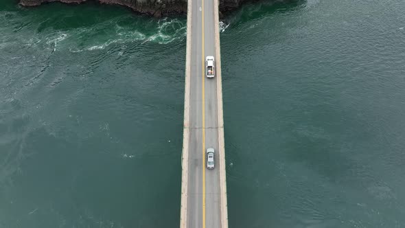 Top down aerial of cars passing over a bridge with vibrant blue water underneath.