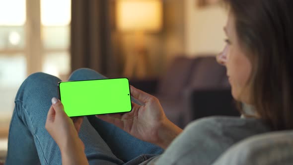 Woman at Home Lying on a Sofa and Using Smartphone with Green Mock-up Screen in Horizontal Mode