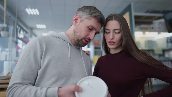 Concentrated Caucasian Couple Talking Looking at Paint Can Standing Between Rows of Goods