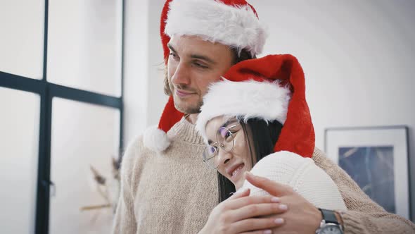 Mixed Race Couple Man in Santa Hat is Smiling Hugging with Love an Asian Woman Standing at Home