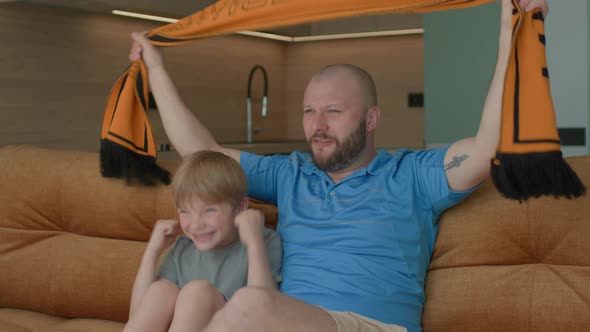 Dad with His Son Watching Football Game Sitting on Couch and Holding Scarf