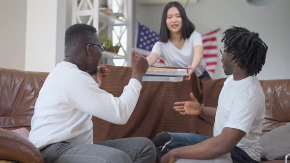 African American Men Talking Sitting on Couch As Asian Woman Bringing Delivered Pizza