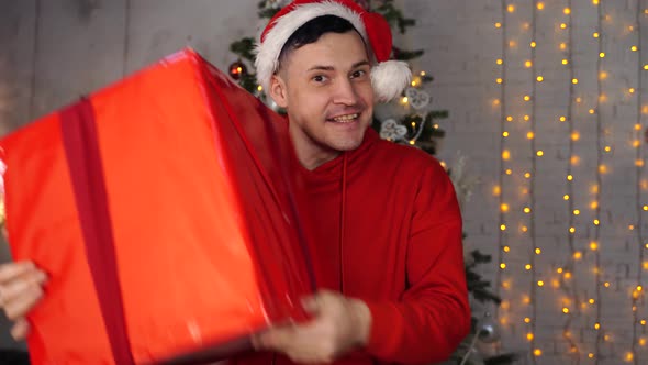 Young Handsome Man in Santa Claus Hat Dancing with Big Gift on Background of Christmas Tree