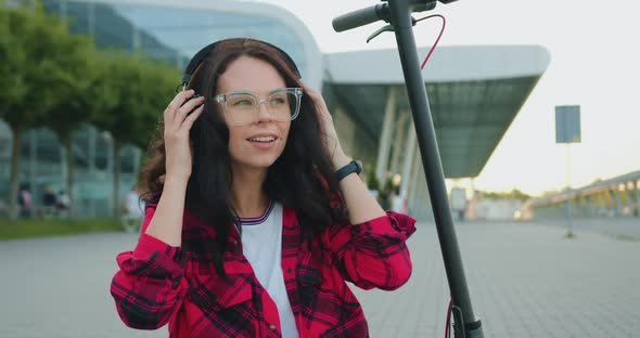 Woman in Stylish Clothes in Glasses Putting on Headphones while Sitting on E-scooter