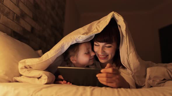 Young Mother and Her Daughter Lying on Bed Under Blanket with Tablet Pc Watching Movie