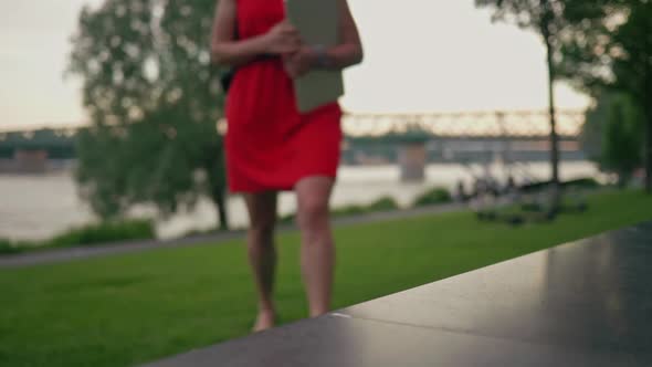Beautiful woman in red dress with a laptop walking towards the bench in the park and sits on it. Cli