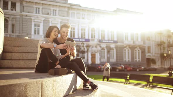 Young Beautiful Couple Smiling Speaking Looking at Tablet Sitting in City Park