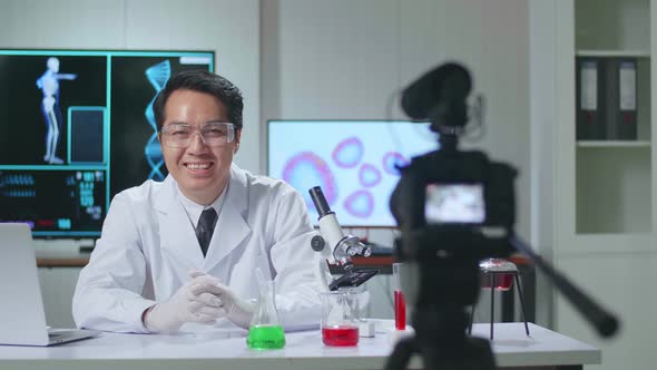 Asian Scientist Researching In The Laboratory With A Microscope, He Warmly Smiles Into The Camera