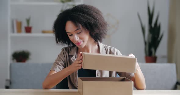 Excited Smiling Curly Mixed Race Woman Receives Gift, Holds Open Cardboard Box, Looks Inside