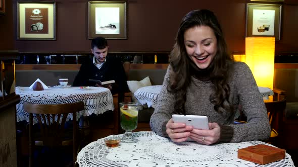 Laughing Girl Having Tea in a Coffee Shop and Boy Reads an Interesting Book Behind Her