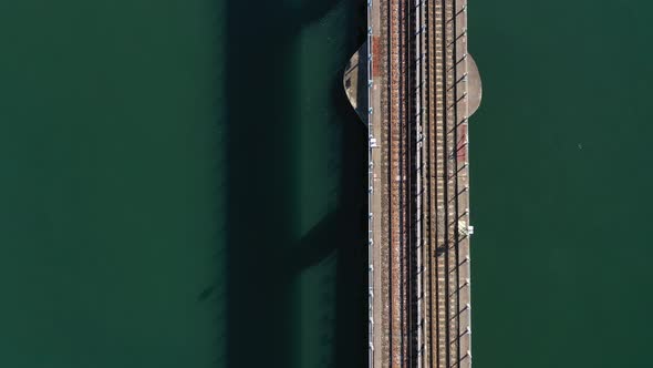 An aerial shot over train tracks over a bay with green water on a sunny day. The drone camera is til